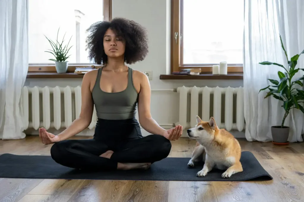 A young woman sitting cross legged on a yoga mat with a dog next to her.