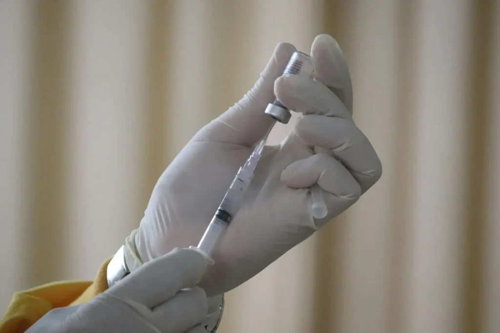 Hands with gloves use a syringe to take medicine out of a vial.